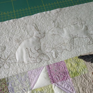 May include: White fabric with embroidered images of two rabbits and floral designs. The fabric is quilted and has a textured surface.