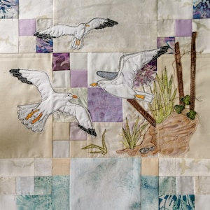 May include: A white quilt with a patchwork design featuring embroidered seagulls and a brown embroidered rock with green plants.