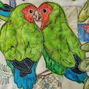 Peut inclure: Deux tourtereaux verts avec des plumes de queue bleues et noires sont cousus ensemble sur un fond de tissu blanc. Les oiseaux se font face, leurs becs se touchant. Les oiseaux sont soulignés d'un fil noir.