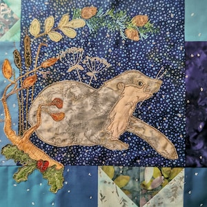 Könnte beinhalten: Ein blauer und weißer Stoff-Patchwork-Quadrat mit einer gestickten Abbildung eines grauen Otters mit einem braunen Schwanz und einem braunen Zweig mit grünen Blättern. Der Hintergrund ist ein blauer Stoff mit weißen Punkten.
