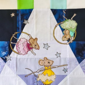 Puede incluir: Una colcha de tela blanca con un diseño de patchwork que presenta tres ratones bordados. Un ratón lleva un vestido rosa y sostiene un aro, otro lleva un vestido amarillo y sostiene un palo, y el tercero lleva un vestido verde y sostiene un aro. La colcha está decorada con estrellas bordadas.