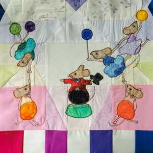 Könnte beinhalten: Ein farbenfroher Quilt mit Patchwork-Design, auf dem gestickte Mäuse verschiedene Zirkusnummern aufführen. Die Mäuse tragen farbenfrohe Kostüme und halten Ballons, einen Hut und ein Seil. Der Quilt besteht aus verschiedenen Stoffen in den Farben Rosa, Lila, Blau, Grün und Weiß.