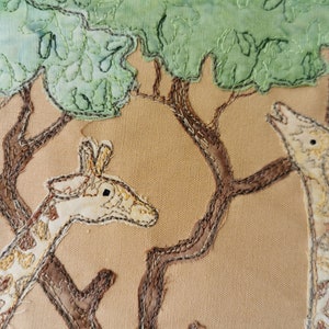 May include: Two embroidered giraffes on a beige fabric background. The giraffes are brown and white with a light brown tree behind them.