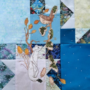 May include: A white fabric square with a white embroidered weasel, a brown embroidered bird, and a green embroidered vine. The background is a patchwork of blue, white, and green fabric squares.
