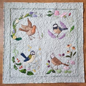 以下が含まれることがあります： 薄い青色のキルティング生地の正方形に、鳥と花の刺繍が施されています。鳥は、ルージュゴージュ、ブルーティット、そして2羽のクレストティットです。花はピンク、パープル、イエローの色合いです。