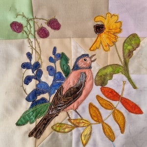 Può includere: Un uccello rosa e marrone con la testa blu è cucito su uno sfondo di tessuto bianco. L'uccello è circondato da fiori e foglie verdi, blu e gialli.