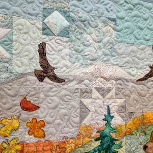 May include: A quilted wall hanging depicting a winter scene with a mountain range, two birds in flight, and a forest with a bear. The background is a patchwork of blue and white fabric.