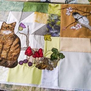 May include: A patchwork quilt with a cat, mushrooms, and a bird embroidered on it. The quilt is made of various fabrics in shades of brown, green, white, and blue.