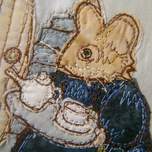 May include: A close-up of a brown and white embroidered mouse holding a white teapot. The mouse is wearing a blue and white jacket.
