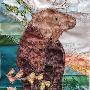 May include: A brown moose with large antlers stands in a green field with yellow and orange flowers. The background is a patchwork of blue, green, and white fabric.