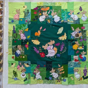 Puede incluir: Una colcha de retazos verde y blanca con ratones y flores bordadas. La colcha está hecha de diferentes cuadrados de tela, cada uno con un diseño diferente. Los ratones llevan todos ropa diferente y están en diferentes poses. Las flores son de varios colores y están dispuestas en un patrón aleatorio.