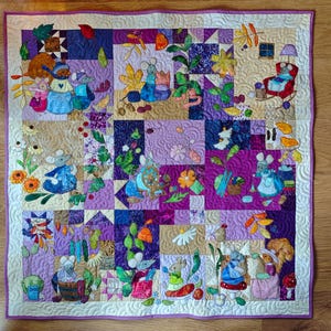 May include: A handmade quilt featuring a patchwork design with various squares in shades of purple, tan, and blue. The quilt is adorned with embroidered images of mice in different scenes, surrounded by flowers and leaves. The quilt has a white border.
