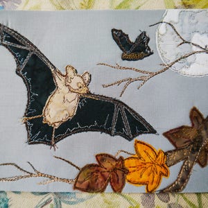 May include: A hand-stitched fabric art piece depicting a bat with brown and black wings, a small butterfly, and autumn leaves on a branch against a gray background with a white moon.