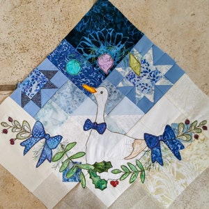 May include: A white goose with a blue bow tie is embroidered on a white fabric square. The goose is surrounded by blue and green embroidered leaves and a red berry. The fabric square is part of a larger quilt with blue and white fabric squares.