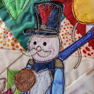 Puede incluir: Arte de tela bordada con un personaje caprichoso con un sombrero azul, barba blanca y un objeto redondo marrón. El personaje está rodeado de piezas de tela coloridas en un diseño de patchwork.