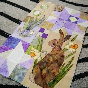 May include: A beige fabric quilt with a patchwork design featuring a brown rabbit, purple and white flowers, and a purple star.