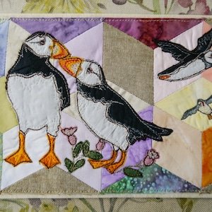 May include: A quilted wall hanging featuring puffins. Two puffins stand side-by-side, with two others in flight. The background is composed of geometric shapes in various colors, including purple, yellow, and orange. The puffins have black and white bodies, orange beaks, and orange feet.