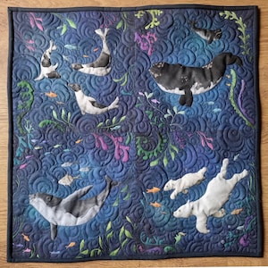 May include: A square, quilted wall hanging with an underwater scene. The dark blue fabric features whales, seals, fish, and seaweed in shades of white, black, orange, green, and purple. The quilting creates a textured, wave-like pattern.