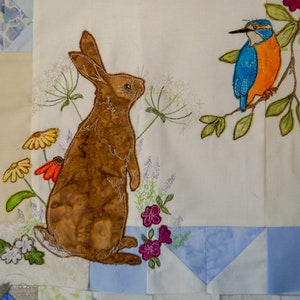 Könnte beinhalten: Eine gestickte Kunstarbeit mit einem braunen Kaninchen, das in einem Blumenfeld steht, und einem blauen und orangefarbenen Eisvogel, der rechts auf einem Ast sitzt.