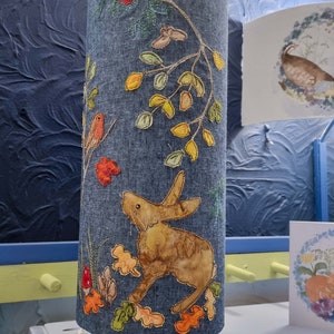 May include: A blue denim lampshade with a brown rabbit, red birds, and yellow and green leaves embroidered on it. The lampshade is on a white metal stand.