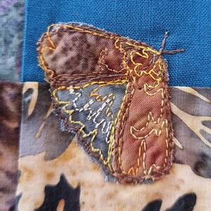 Op de afbeelding: Een bruine en grijze stoffen vlinder met gele stiksels, genaaid op een patchwork stoffen achtergrond.
