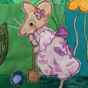 Könnte beinhalten: Eine braune Maus, die ein rosa Kleid mit Blumenmuster und eine rosa Blume auf dem Kopf trägt. Die Maus hält eine kleine braune Schaufel und steht auf grünem Hintergrund.
