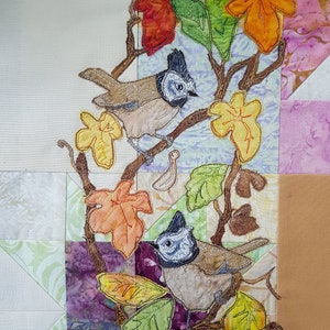 Könnte beinhalten: Zwei braune und graue Vögel mit blauen und weißen Markierungen sitzen auf einem Ast mit orangefarbenen, gelben und grünen Blättern. Die Vögel sind von einem Patchwork aus Stoff in verschiedenen Farben und Mustern umgeben.