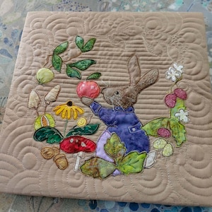 Puede incluir: Un cuadrado de tela beige acolchado con un conejo marrón que sostiene una manzana roja. El conejo está rodeado de hojas verdes, flores amarillas y setas marrones. La tela tiene un patrón acolchado de líneas onduladas.