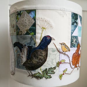 May include: A white lampshade with a patchwork design featuring embroidered birds, flowers, and leaves. The birds are a black grouse and a small yellow bird. The flowers are white and the leaves are green.