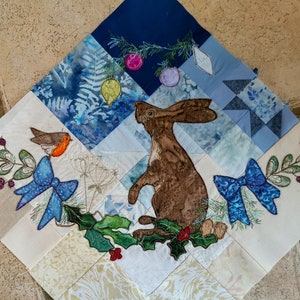 May include: A white fabric square with a brown rabbit, a red robin, and blue bows embroidered on it. The fabric is divided into smaller squares with a blue and white pattern.