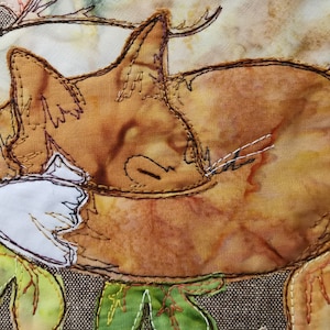 Puede incluir: Primer plano de una aplicación de tela de gato marrón y naranja con detalles en tela blanca, cosida sobre un fondo de tela beige. El gato está enroscado en una posición de dormir con los ojos cerrados.