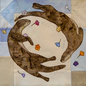 Könnte beinhalten: Zwei braune Otter schwimmen in einem Kreis, umgeben von bunten Blumen. Die Otter sind aus Stoff und auf einen weißen Hintergrund genäht.