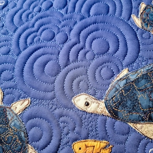 Puede incluir: Primer plano de una tela acolchada azul con tortugas marinas bordadas. Las tortugas tienen caparazones estampados en beige y azul, con cabezas y aletas beige. El fondo presenta un patrón arremolinado, dando la impresión de agua.