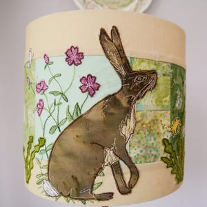 May include: A cylindrical lampshade featuring a detailed appliqué design. The design includes a brown hare, pink flowers, green foliage, and a butterfly against a light green and beige background. The lampshade is a decorative home decor item.