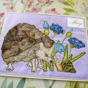 May include: A purple fabric card with a brown hedgehog, a green snail, and blue flowers embroidered on it. The card has the word "May" written on it.