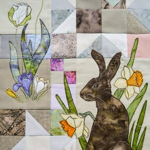 Puede incluir: Un conejo marrón bordado sobre un fondo de tela beige. El conejo está rodeado de flores verdes y blancas. El fondo es un mosaico de diferentes telas en varios tonos de beige, blanco y gris.
