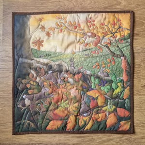 May include: A quilted wall hanging showing an autumnal landscape. The artwork features a tree with orange and red leaves, squirrels, and rabbits amongst fallen leaves. The colour palette includes shades of brown, green, orange, and yellow.