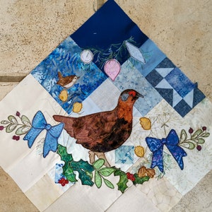Peut inclure: Un carré de tissu blanc avec un oiseau brun et blanc, des nœuds bleus et des feuilles de houx vertes brodés dessus. Le tissu est divisé en carrés plus petits avec différents motifs bleus.