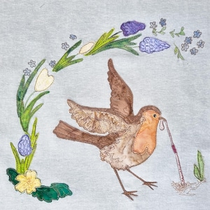 May include: A brown and orange robin with a worm in its beak, surrounded by a wreath of green leaves, yellow flowers, and purple blossoms.