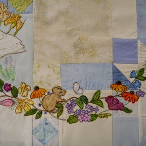May include: A white swan embroidered on a patchwork quilt with a brown mouse, flowers, and leaves. The quilt is made of various shades of blue and white fabric.