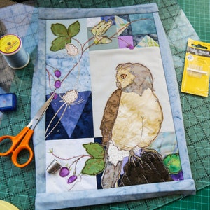 Op de afbeelding: Een blauwe en witte patchwork quiltblok met een geborduurde uil en bloemmotieven. De uil is bruin, wit en beige met een lichtbruine snavel. De bloemen zijn wit en roze met groene bladeren.