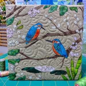Op de afbeelding: Een gewatteerd kunstwerk van stof met twee blauw-oranje ijsvogels op bruine takken. Witte bloemen en groene bladeren sieren de beige achtergrond, die een gewatteerd patroon heeft.