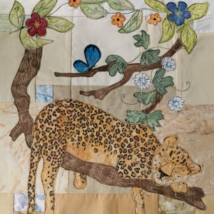 May include: A brown and black leopard sleeping on a branch with green leaves and blue and white flowers. The leopard is embroidered on a white fabric background.