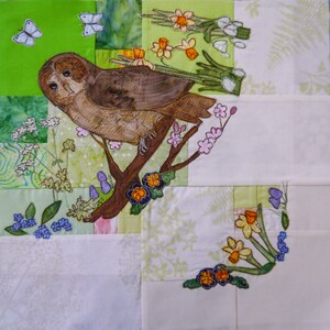 Könnte beinhalten: Eine braune Eule sitzt auf einem Ast mit rosa und violetten Blüten. Die Eule ist von grünen Blättern und weißen Blüten umgeben. Der Hintergrund ist ein Patchwork aus grünem, weißem und braunem Stoff.