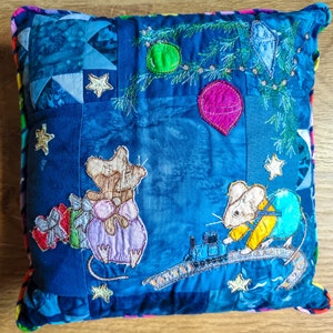 May include: A blue and white patchwork pillow with embroidered mice, a train, and Christmas ornaments. The mice are holding presents and the train is on a track. The pillow is decorated with stars and a pine tree.