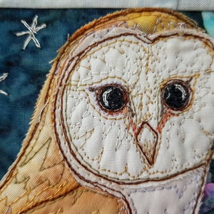 May include: Close-up of a white barn owl with brown and yellow feathers, embroidered on a blue fabric background. The owl's eyes are black and the beak is orange.