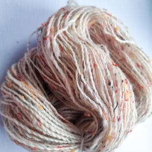 May include: A skein of yarn in a neutral color with flecks of red, yellow, and black. The yarn is tightly wound, showing the texture and variations in the fibers. Ideal for knitting or crocheting projects.