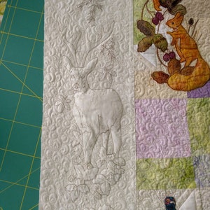 May include: A white fabric panel with an embroidered deer with antlers, surrounded by leaves and vines. The deer is outlined in brown thread. The fabric has a textured background.