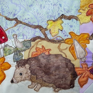 Könnte beinhalten: Ein brauner Igel mit weißem Bauch und schwarzen Augen liegt in einem Haufen Herbstlaub. Die Blätter sind orange, gelb und rot. Es gibt auch Pilze und Beeren in der Szene.