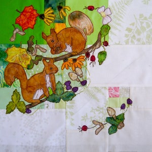 Könnte beinhalten: Ein weißer Stoff-Patchworkblock mit einem gestickten Design aus zwei roten Eichhörnchen auf einem Ast mit Blättern, Beeren und Pilzen. Die Eichhörnchen sind braun mit weißen Akzenten.